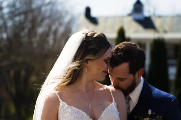 A bride in a white gown and veil and a groom in a suit share a close moment outdoors with a building in the background.