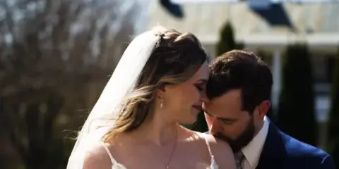 A bride in a white gown and veil and a groom in a suit share a close moment outdoors with a building in the background.