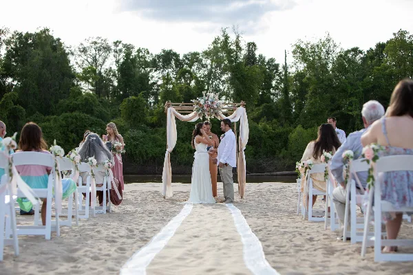 A wedding ceremony in progress on a sandy river beach with a floral arch and seated guests.