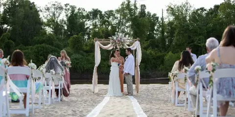 A wedding ceremony in progress on a sandy river beach with a floral arch and seated guests.