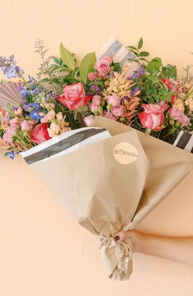 A hand holding a wrapped bouquet of pink roses and mixed flowers with a Blossom logo sticker on the kraft paper wrapping.