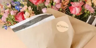 A hand holding a wrapped bouquet of pink roses and mixed flowers with a Blossom logo sticker on the kraft paper wrapping.