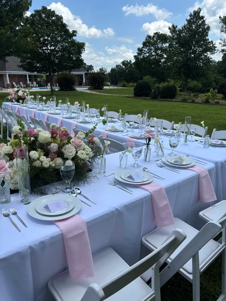 An outdoor wedding reception table set with white linens, pink accents, floral arrangements, and place settings.