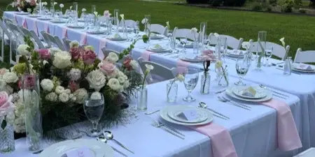 An outdoor wedding reception table set with white linens, pink accents, floral arrangements, and place settings.