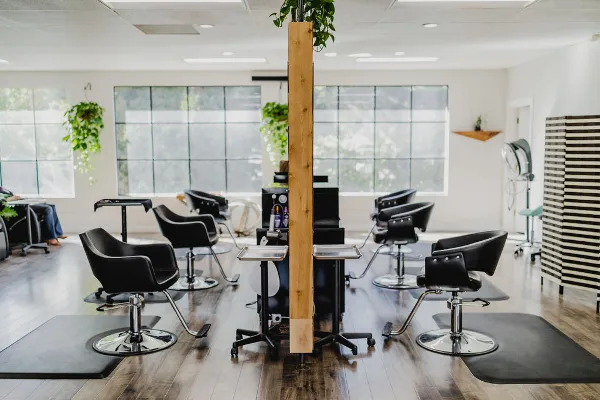 Interior view of a modern hair salon featuring black styling chairs, mirrors, and a central wooden pillar with plants.