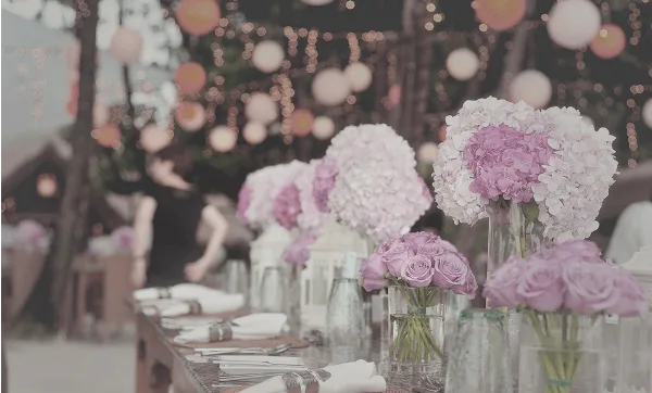 A long outdoor event table set with white and pink floral centerpieces, glassware, and string lights in the background.