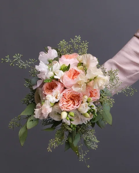 A hand-held bridal bouquet composed of peach and cream garden roses, ranunculus, and greenery against a neutral grey background.