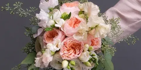 A hand-held bridal bouquet composed of peach and cream garden roses, ranunculus, and greenery against a neutral grey background.
