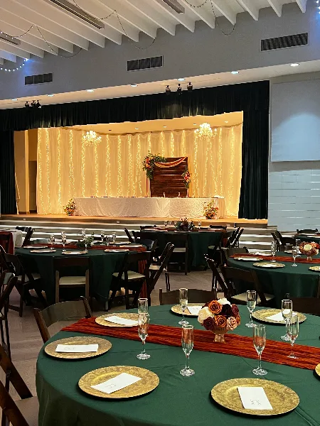 Wedding reception setup in a banquet hall with green tablecloths, gold chargers, floral centerpieces, and a decorated head table under chandeliers.