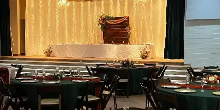Wedding reception setup in a banquet hall with green tablecloths, gold chargers, floral centerpieces, and a decorated head table under chandeliers.