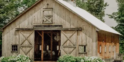 Exterior view of a rustic wooden barn with a white roof and cupola, featuring open sliding doors and white hydrangea bushes in the foreground.