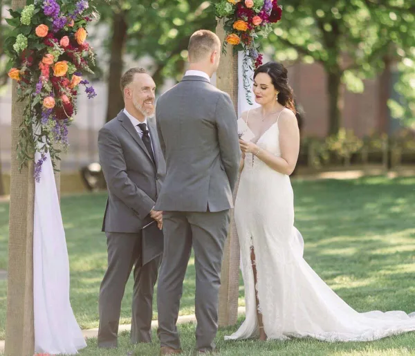 A bride and groom stand before an officiant at a floral-decorated wedding arch outdoors.