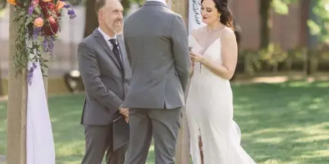 A bride and groom stand before an officiant at a floral-decorated wedding arch outdoors.