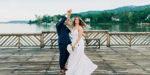 A bride and groom dancing on a wooden dock with a lake and hills in the background.