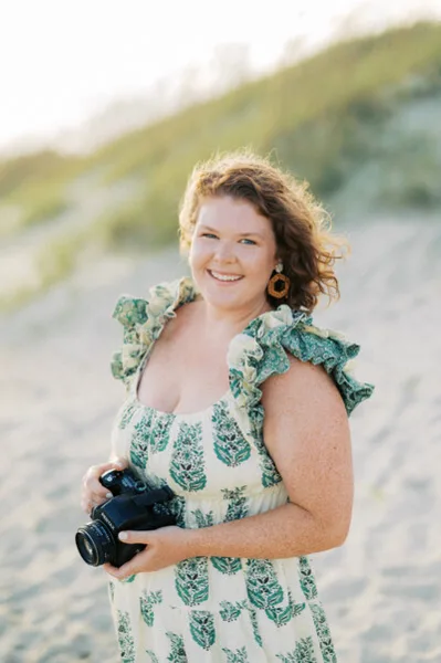 A smiling woman holding a black camera on a sandy beach.