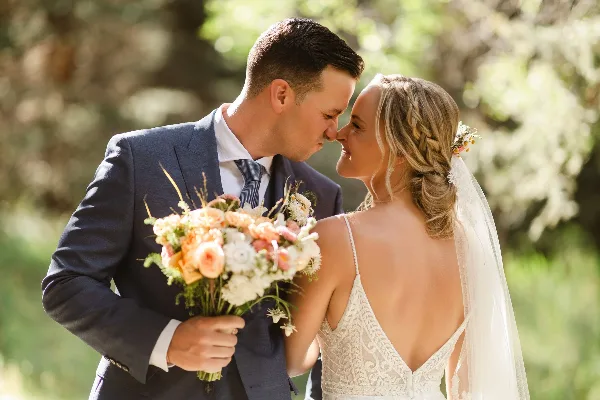 Bride and groom in wedding attire sharing a close moment outdoors with a floral bouquet.