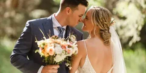 Bride and groom in wedding attire sharing a close moment outdoors with a floral bouquet.