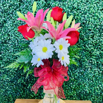 A floral arrangement containing red roses, pink lilies, and white daisies tied with a red ribbon, set against a green leafy background.