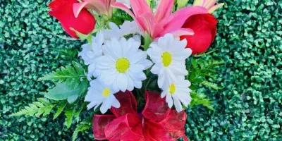 A floral arrangement containing red roses, pink lilies, and white daisies tied with a red ribbon, set against a green leafy background.