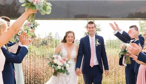 A bride and groom walking down the aisle holding hands, surrounded by a wedding party clapping and holding bouquets.