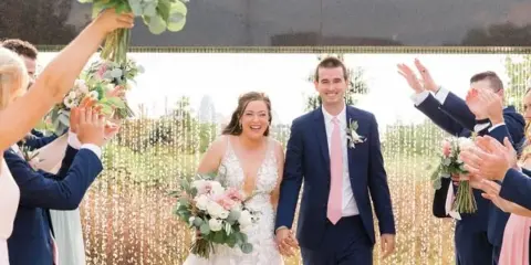 A bride and groom walking down the aisle holding hands, surrounded by a wedding party clapping and holding bouquets.