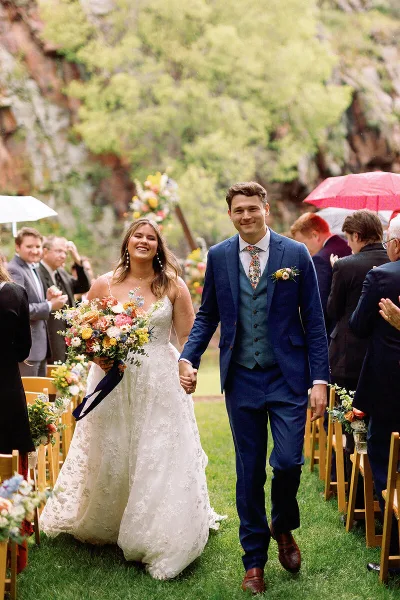 A bride and groom walking down the aisle at an outdoor wedding ceremony, holding hands and smiling.