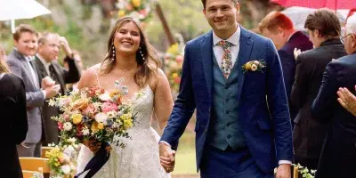 A bride and groom walking down the aisle at an outdoor wedding ceremony, holding hands and smiling.