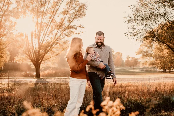 A family of three posing outdoors in a field during golden hour, with the father holding a baby and the mother standing beside them.