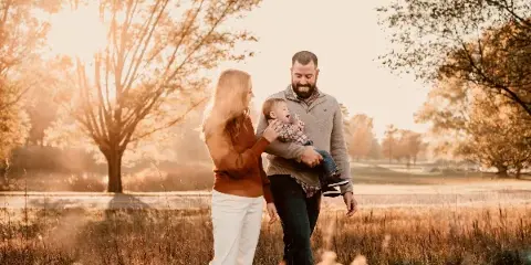 A family of three posing outdoors in a field during golden hour, with the father holding a baby and the mother standing beside them.