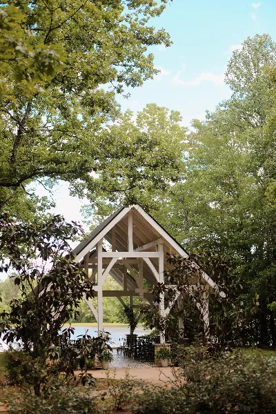 A white timber-frame wedding chapel with a peaked roof, set among trees with a lake visible in the background.