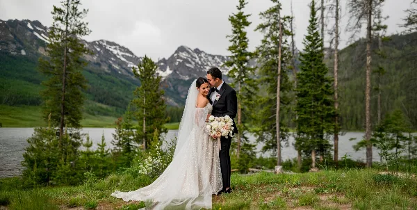 A bride and groom standing together in a mountain landscape with a lake and pine trees in the background.
