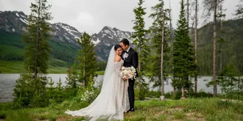 A bride and groom standing together in a mountain landscape with a lake and pine trees in the background.