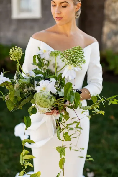A bride in a white off-the-shoulder gown holding a lush bouquet of white flowers and greenery.