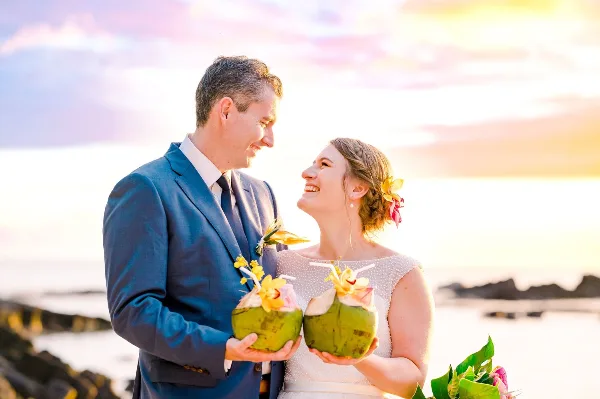 A bride and groom smiling at each other while holding fresh coconuts on a beach at sunset.
