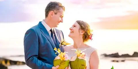 A bride and groom smiling at each other while holding fresh coconuts on a beach at sunset.