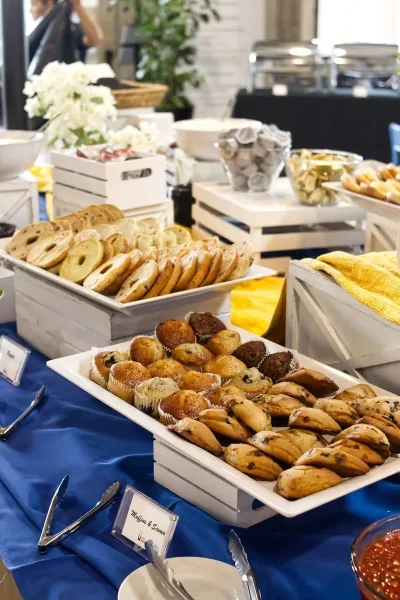 A catering buffet table featuring trays of bagels, muffins, and pastries with serving tongs.