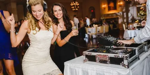 A professional DJ operating a turntable setup at a wedding reception while a guest dances nearby.