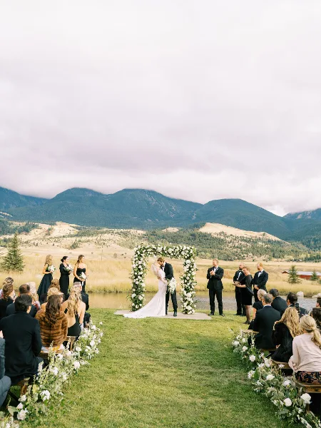 Outdoor wedding ceremony with a floral arch, wedding party, and guests in a Montana mountain setting.