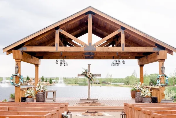 A rustic wooden wedding pavilion with a cross altar, floral arrangements, and a lake view in the background.