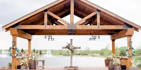 A rustic wooden wedding pavilion with a cross altar, floral arrangements, and a lake view in the background.