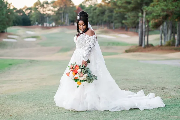 A bride in a white lace wedding gown and veil stands on a golf course holding a colorful bouquet.