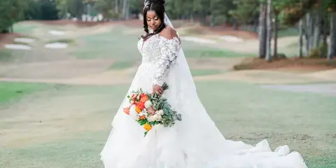 A bride in a white lace wedding gown and veil stands on a golf course holding a colorful bouquet.