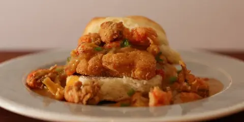 A biscuit sandwich topped with fried shrimp and gravy on a white plate.