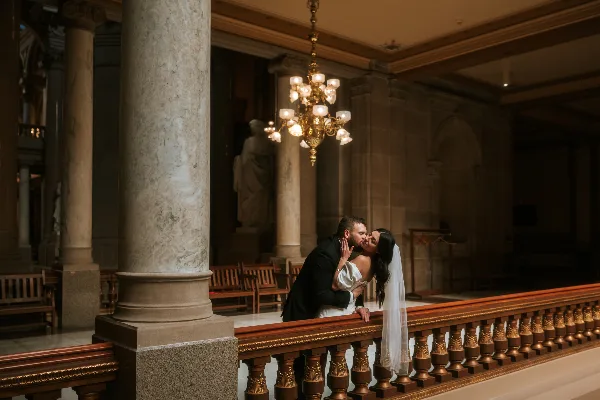 A bride and groom sharing a kiss in a grand, ornate interior with marble columns and a chandelier.