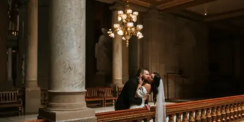 A bride and groom sharing a kiss in a grand, ornate interior with marble columns and a chandelier.