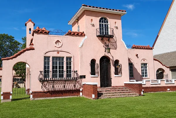 A pink historic house with a red tile roof and wrought iron balconies, set on a green lawn under a blue sky.