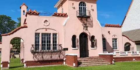 A pink historic house with a red tile roof and wrought iron balconies, set on a green lawn under a blue sky.