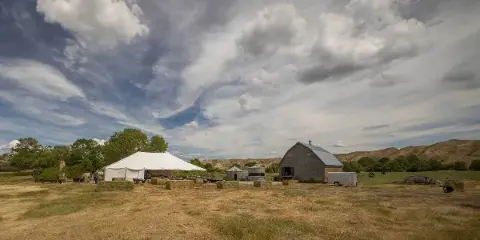 A wide shot of a rustic wooden barn with a blue roof and a white event tent set up on a grassy ranch field under a cloudy sky.