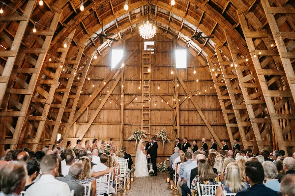 A wedding ceremony in progress inside a rustic wooden barn with a high vaulted ceiling, string lights, and a chandelier, surrounded by seated guests.