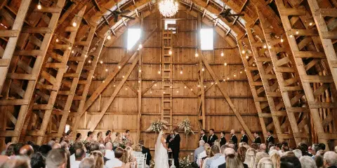 A wedding ceremony in progress inside a rustic wooden barn with a high vaulted ceiling, string lights, and a chandelier, surrounded by seated guests.